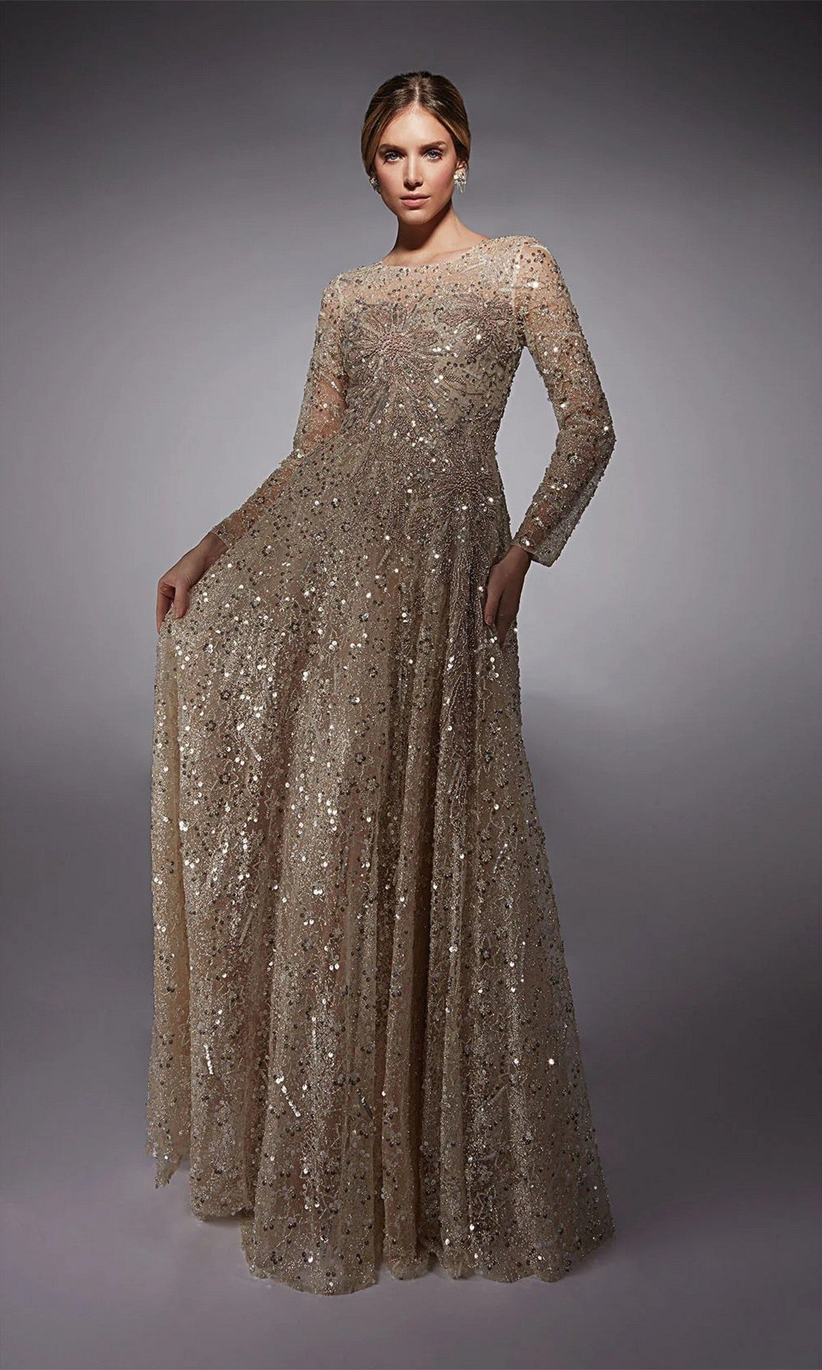 A woman wears the Alyce 27757 long formal dress—full-length, long-sleeved, and covered in gold sequins—making it a chic mother-of-the-bride look against a plain gray background.